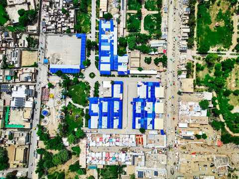 Aerial view of the Model Bazar featuring organized rows of blue-roofed market stalls, surrounding urban infrastructure, and green spaces in Dera Ghazi Khan, Punjab, Pakistan.