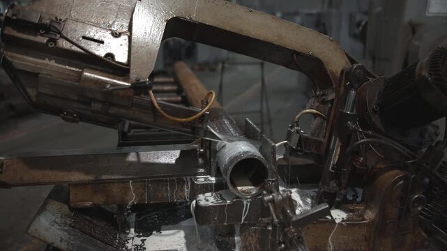 Industrial bandsaw cutting steel pipe with coolant spray and metal shavings inside dim workshop, heavy machine clamped in vise, showing fabrication process with wet sparks and grime