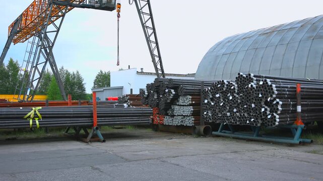 Gantry crane with hanging chain hook above organized stacks of bundled steel pipes beside curved corrugated warehouse wall in outdoor industrial storage yard, with industrial, pipe, steel, crane