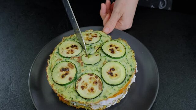 Person cooking a layered zucchini tart with pesto on a dark counter, using a silver knife to cut the savory dish in a Cooking setting.