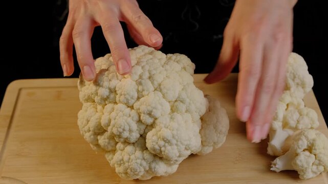 Adult hands in dark clothing examine a large head of white cauliflower and a cut piece on a wooden cutting board in a cooking studio with black background.