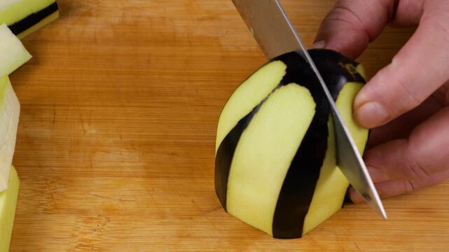 A chef uses a sharp knife to cut a striped peeled eggplant into pieces on a wooden cutting board, preparing healthy organic ingredients with visible flesh and skin textures.