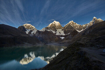 Obraz premium Beneath a dramatic sky, the snow crowned summits of Siula, Yerupajá, and Jirishanca rise in perfect formation above Carhuacocha Lagoon, their jagged silhouettes mirrored flawlessly in the still, glass