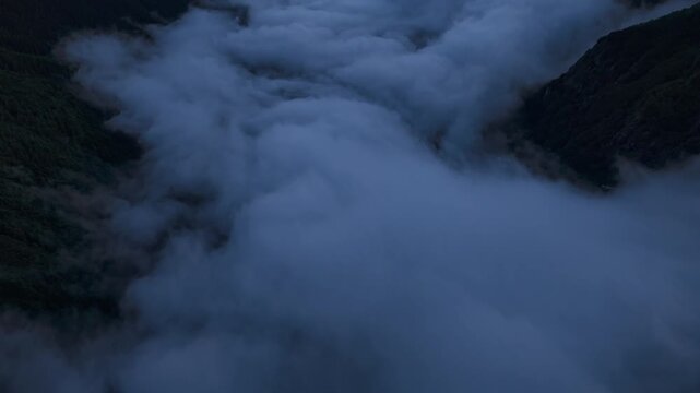 A dramatic aerial perspective showing dense fog and low clouds flowing through a rugged mountain valley in Valseriana during twilight. Suitable for nature and travel documentaries.