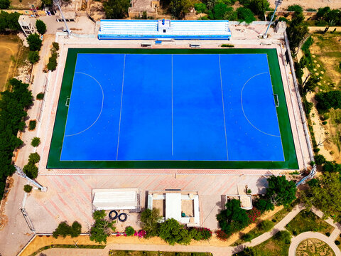 Aerial view of the hockey stadium with a blue pitch and green borders surrounded by sports infrastructure and trees in Dera Ghazi Khan, Punjab, Pakistan.