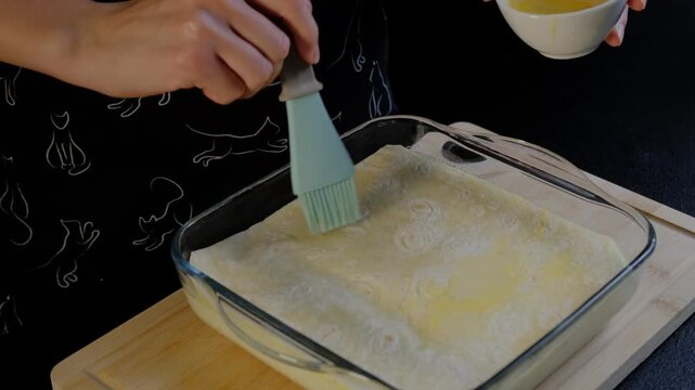 Adult hands use a pastry brush to spread melted butter over dough in a glass baking dish on a wooden cutting board in a Cooking kitchen.