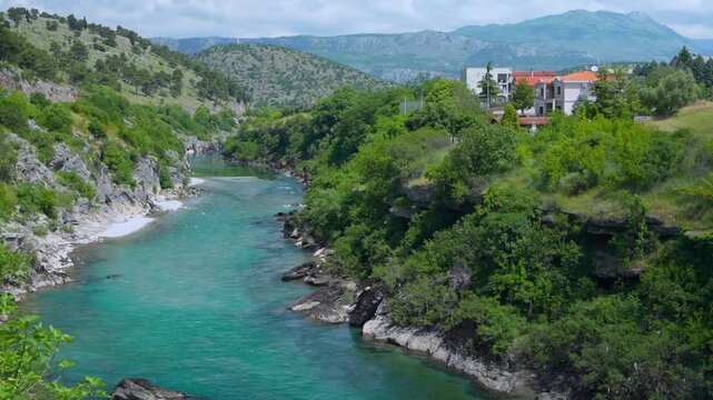A turquoise river flows through a rocky canyon lined with green trees near hillside houses, with mountains rising in the background near Podgorica Montenegro.