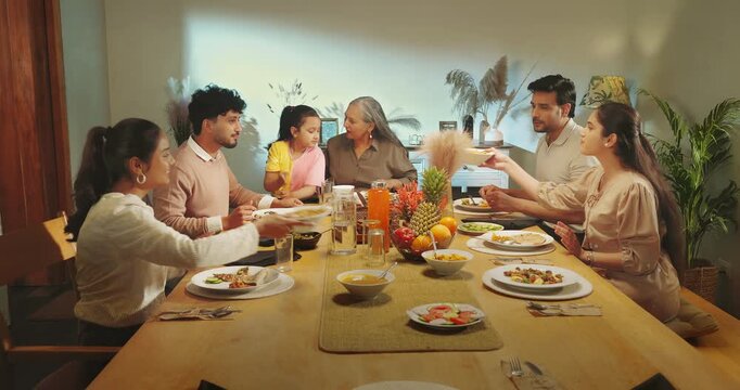 Indian happy joint family group sitting around table eat food talk speak share meal hold jug pour water into glass young adult male female mom girl boy child spend time enjoy night room home place