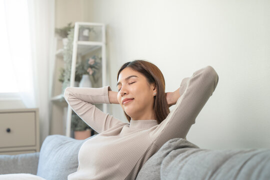 Happy relaxed young Asian woman resting on a comfortable sofa with hands behind head, enjoying peaceful weekend leisure time and stress-free breathing at home.