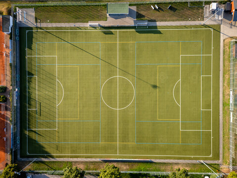 Aerial view of a green artificial turf football pitch with white and yellow markings, surrounded by fencing and trees in Germany.