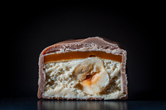 Macro closeup of milk chocolate candy bar cut half showing textures of chewy caramel layer soft nougat cream and whole crunchy hazelnut inside on dark background.