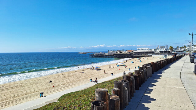 The sandy Redondo Beach shore in California, showing sunbathers, a paved bicycle path, wooden barrier posts, and the historic pier under a clear blue sky. USA