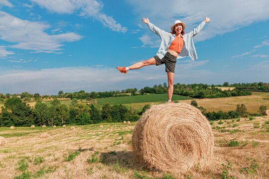 Exuberant young woman kicks one leg high atop a round hay bale, arms flung wide, laughing freely across a rolling green countryside. Adventure concept.