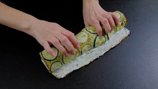 Adult hands roll sliced zucchini onto a bread loaf spread with white substance on a dark countertop in a kitchen, Cooking location, daytime.