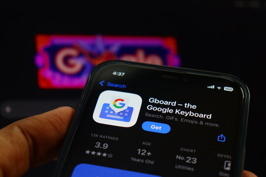 Close-up of a person's hand holding a smartphone displaying the Gboard Google Keyboard application on the Apple iOS App Store, featuring a colorful blurred Google logo in the background.