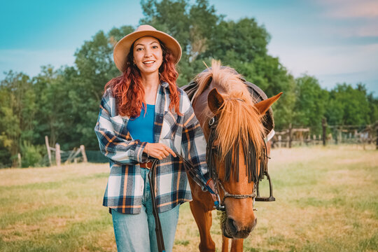 Happy woman wearing a hat, smiling while leading a horse in a grassy field on a countryside ranch