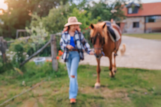 Blurred woman in a straw hat leads a saddled chestnut horse across a sunny farm yard, walking ahead with a relaxed smile. Rural lifestyle concept.