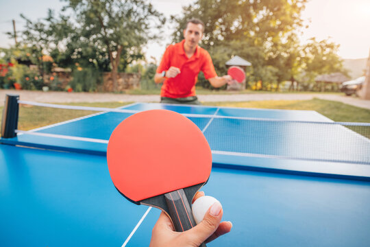 POV first-person perspective of two players competing in an outdoor table tennis match on a sunny afternoon, opponent visible across the net. Active lifestyle concept.