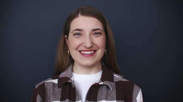 Portrait of smiling young Caucasian woman looking at camera against dark background. Female wearing checkered shirt and hoop earrings. Friendly confident expression for professional profile.