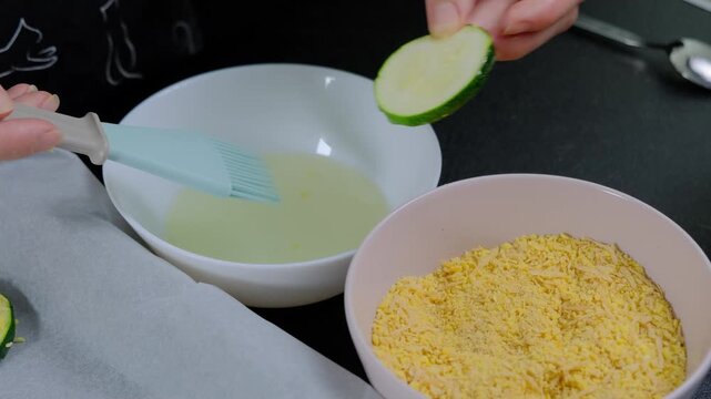 Hands dip zucchini slices in batter, then coat with breadcrumbs on a dark kitchen countertop during daytime cooking.