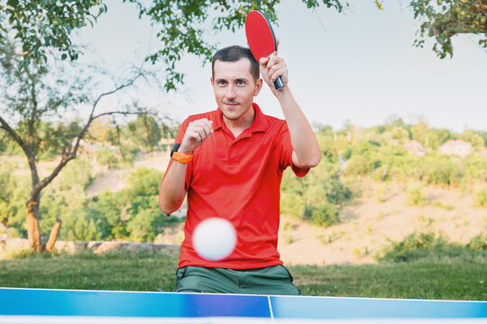 Young man in a red shirt raises a ping pong paddle mid-game at an outdoor tennis table, ball blurred in motion. Active leisure concept.