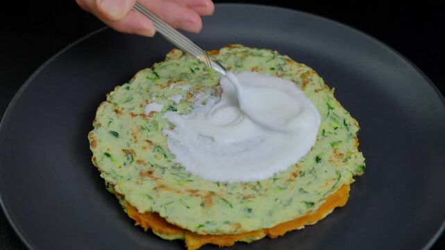 Hands in a kitchen cooking green vegetable fritters, stacking them on a plate and adding white cream with a spoon, at Cooking location.