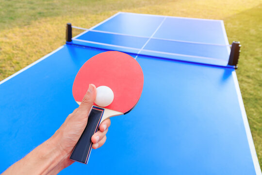 Warm sunlit hand holding a red ping pong paddle with ball over a bright blue outdoor table tennis, ready to serve.