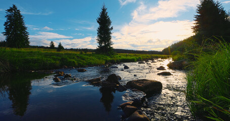 Obraz premium Crystal-clear mountain stream flowing through a peaceful meadow at sunset, reflecting the colorful sky with evergreen trees, rocks and warm golden light in a tranquil natural landscape.