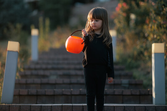 Cute Girl Wearing Black Costume Holding Pumpkin Bucket On Stairs. Excited child stands outside steps ready for autumn Halloween trick or treat walk