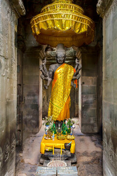 Ancient stone statue of multi-armed Hindu deity draped in gold cloth inside Angkor Wat temple. Altar with offerings, incense and golden ceremonial umbrella. Religion and spirituality concept.