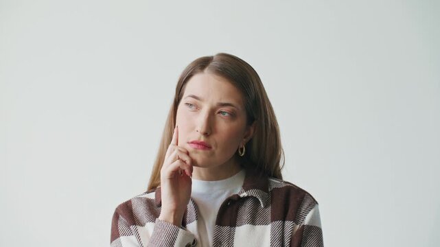 Thoughtful young Caucasian woman resting chin on hand and looking aside in bright studio. Serious female reflecting on decision and analyzing ideas. Calm expression with copy space.