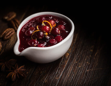 Vibrant homemade cranberry sauce with fresh orange zest in a white serving dish displayed on a dark rustic wooden table with pecans and star anise, perfect for holiday gatherings