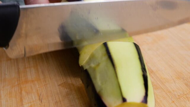 A chef slices a partially peeled eggplant on a wooden cutting board, dicing the fresh vegetable for a healthy meal with precise knife action in a home or restaurant kitchen.