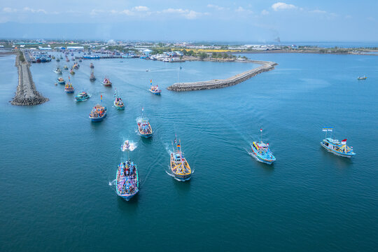 Aerial view of colorful fishing boats sailing in a procession through the Mayangan harbor with calm blue water and a breakwater in Probolinggo, East Java, Indonesia.