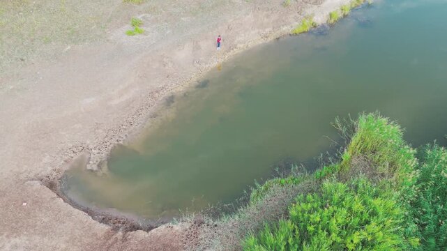 High angle top down 4K drone footage flying over a river bank with green reeds, shallow water, and a tiny silhouette of a person in red walking along the shore. Minimalist natural landscape view in Ce