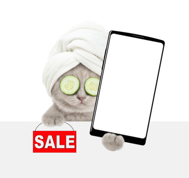Cute tiny kitten wearing towel on it head and slices of cucumer on it eyes holds big smartphone with white blank screen in it paw above blank white banner. isolated on white background