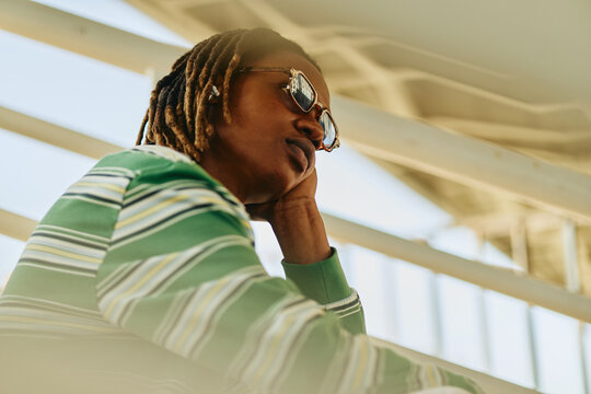 Young adult African American man with dreadlocks sitting outdoors, looking away thoughtfully. Image represents mental wellbeing and youth lifestyle, suitable for mental health campaigns