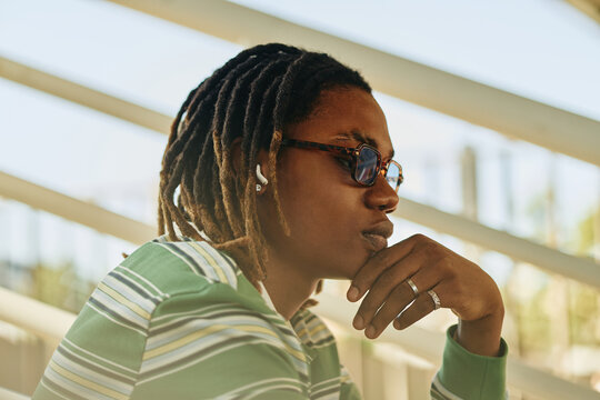 Young adult African American man with dreadlocks wearing sunglasses and wireless earbuds sitting outdoors, thinking. Image suitable for youth lifestyle and modern technology marketing