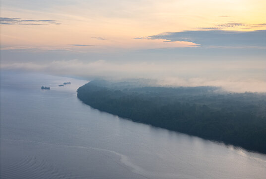 Aerial view of the Kumai River winding through dense tropical rainforest with morning mist hovering over the water and small ships in the distance, Kumai, Central Kalimantan, Indonesia.