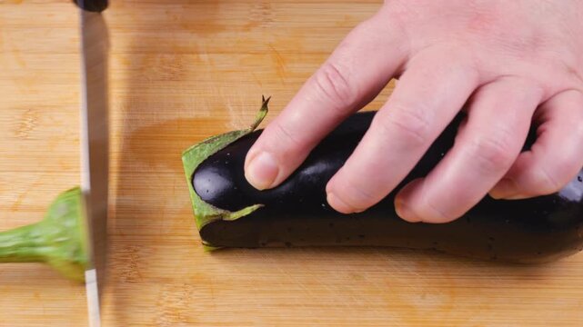 A female chef uses a knife to cut off the stem of a fresh dark purple eggplant on a wooden board, preparing organic raw ingredients for a healthy vegetarian meal.