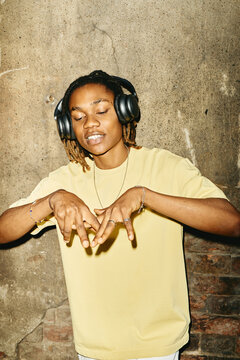 Vertical image of young Black man wearing headphones, dancing outdoors against concrete wall, representing youth culture and music streaming services in modern advertising