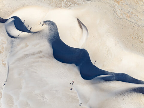 Aerial view of Lancelin Sand Dunes with white sand and dark shadows, featuring small figures of people walking across the undulating landscape in Lancelin, Western Australia, Australia.