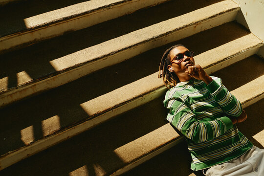 Young adult African American man lying on outdoor concrete stairs, wearing sunglasses and thinking. Image represents youth lifestyle and mental wellbeing, suitable for Gen Z marketing campaigns