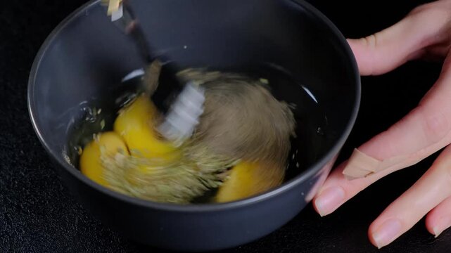 A person whisks egg yolks and powder in a black mixing bowl on a kitchen counter at Cooking, creating a frothy yellow mixture.
