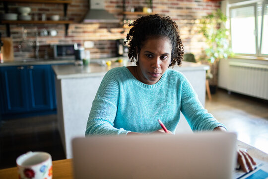 Focused woman taking notes while working on laptop at home kitchen
