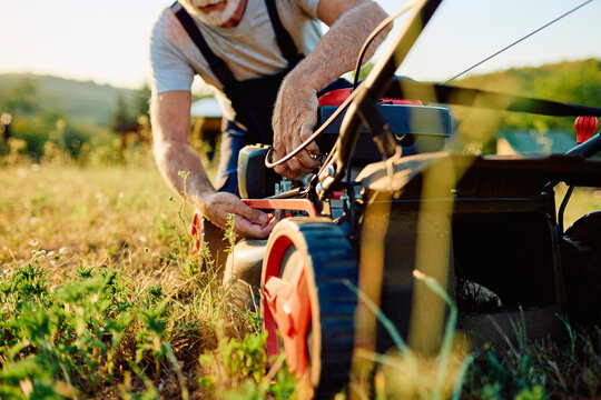 Senior man working on a lawn mower engine in a grassy field during sunset. Focusing on yard work, maintenance, and repair