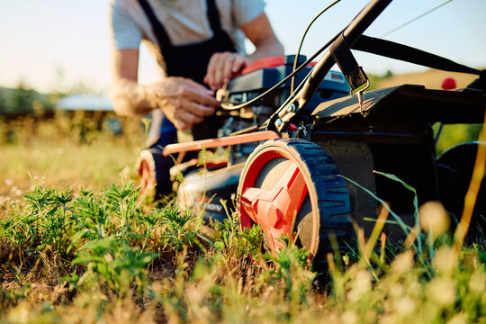 Man adjusting a gas lawn mower engine, preparing to cut overgrown grass in a sunny outdoor yard