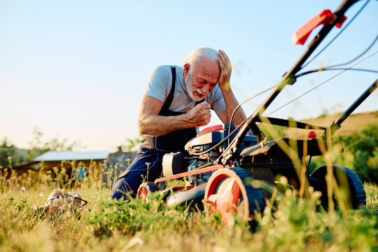 Senior man feeling frustrated and stressed, kneeling beside a broken lawn mower while trying to fix it in his backyard