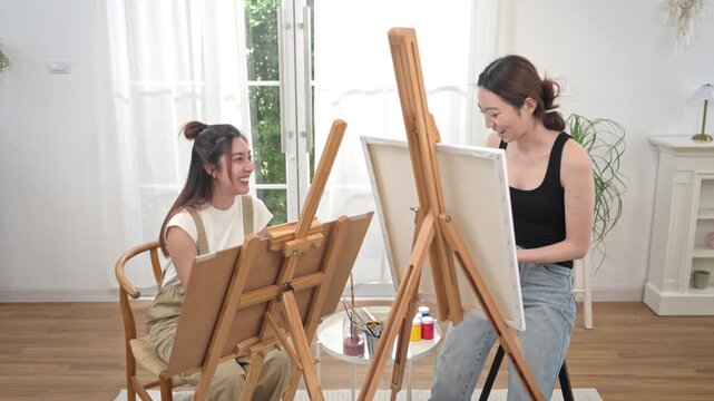 Two young Asian women painting a colorful landscape on canvas together during a relaxing weekend at home, enjoying creative art as a hobby in a bright living room