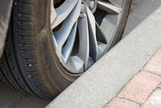 Alloy wheel rim almost contacting sidewalk curb, high risk of severe rim damage and scratching. Car alloy wheel rim pressed against sharp concrete curb edge, tire sidewall compression. Selective focus